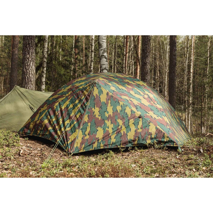 Tienda militar belga antidesgarro impermeable para dos personas instalada en un bosque con diseño de camuflaje y estacas aseguradas.