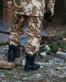 Rear view of person wearing desert camouflage British army combat trousers and black boots near a campfire outdoors
