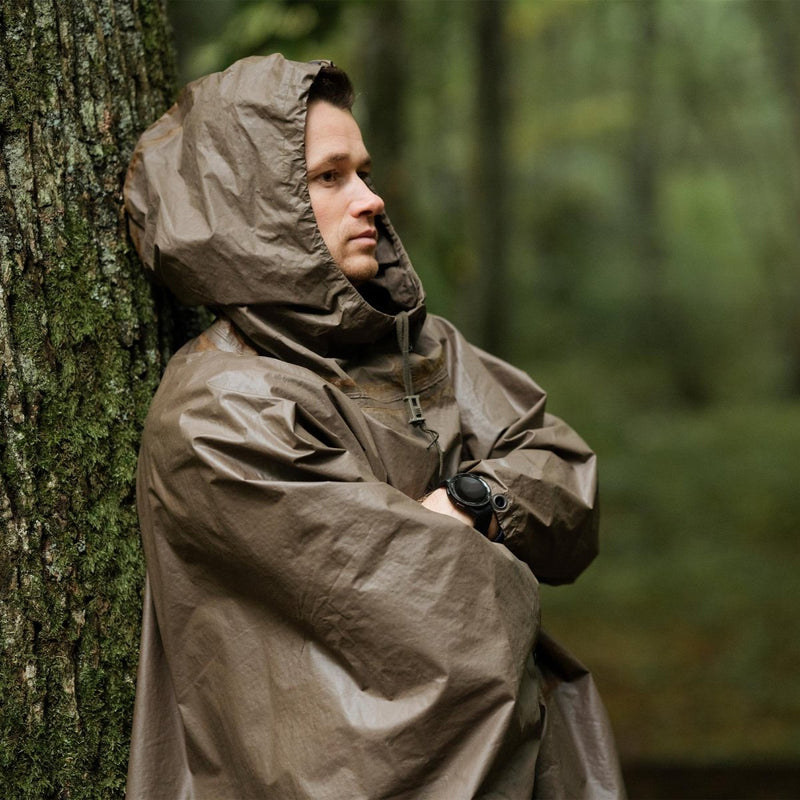 Person wearing vintage German army waterproof rain poncho with hood leaning against tree in forest setting