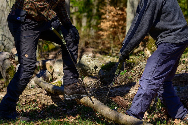 Two people using a Nordic Pocket Saw X-Long edition to cut a thick branch outdoors.